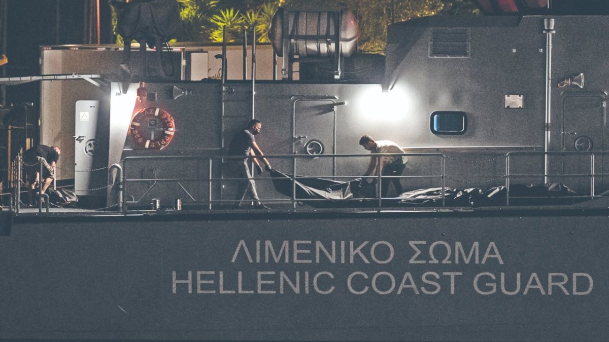 Varios hombres transportaban bolsas con cadáveres desde un buque, tras el naufragio de una embarcación, mientras los sobrevivientes esperaban ayer dentro de un almacén en el puerto de la ciudad de Kalamata