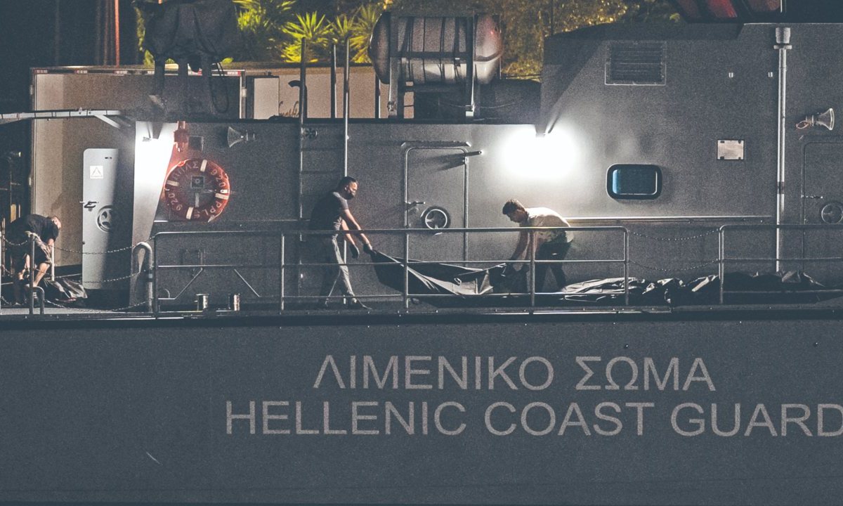 Varios hombres transportaban bolsas con cadáveres desde un buque, tras el naufragio de una embarcación, mientras los sobrevivientes esperaban ayer dentro de un almacén en el puerto de la ciudad de Kalamata